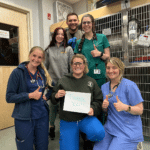 Six veterinary professionals are wearing scrubs and smiling towards the camera. Three of the people in the image are giving a "thumbs up" and the person in the front of the group holds a piece of paper that shows a "Thank You" message.