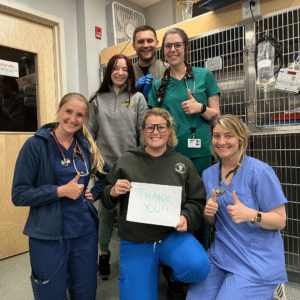 Six veterinary professionals are wearing scrubs and smiling towards the camera. Three of the people in the image are giving a "thumbs up" and the person in the front of the group holds a piece of paper that shows a "Thank You" message.